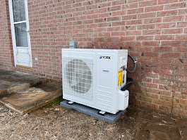 Outdoor heat pump unit installed on a concrete pad against a brick wall of a house.